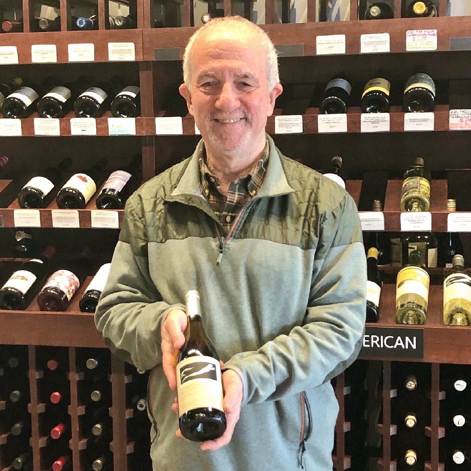 Howie Rubin holding bottle of wine in front of wine racks at Vin Fromage