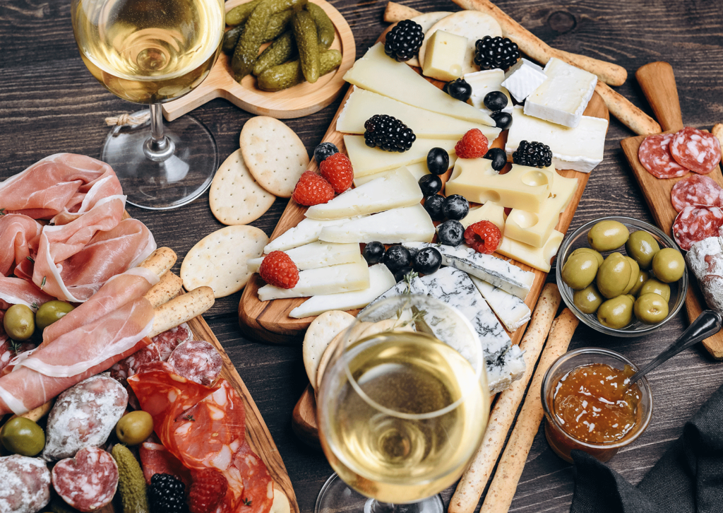 cheese and charcuterie boards with fruits, breads, crackers, jams, olives, gherkins and two glasses of white wine